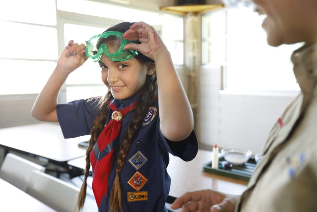 A girl with double braids in a Scouts BSA uniform smiles as she puts on lab goggles, ready to do a science experiment.
