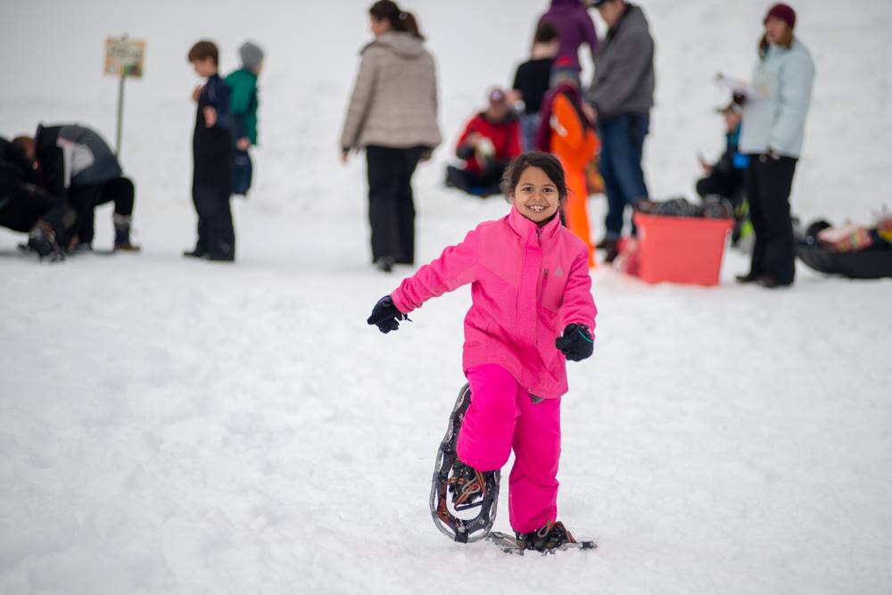 A young girl dressed in a bright pink snow suit smiles as she snow-shoes her way toward the camera.