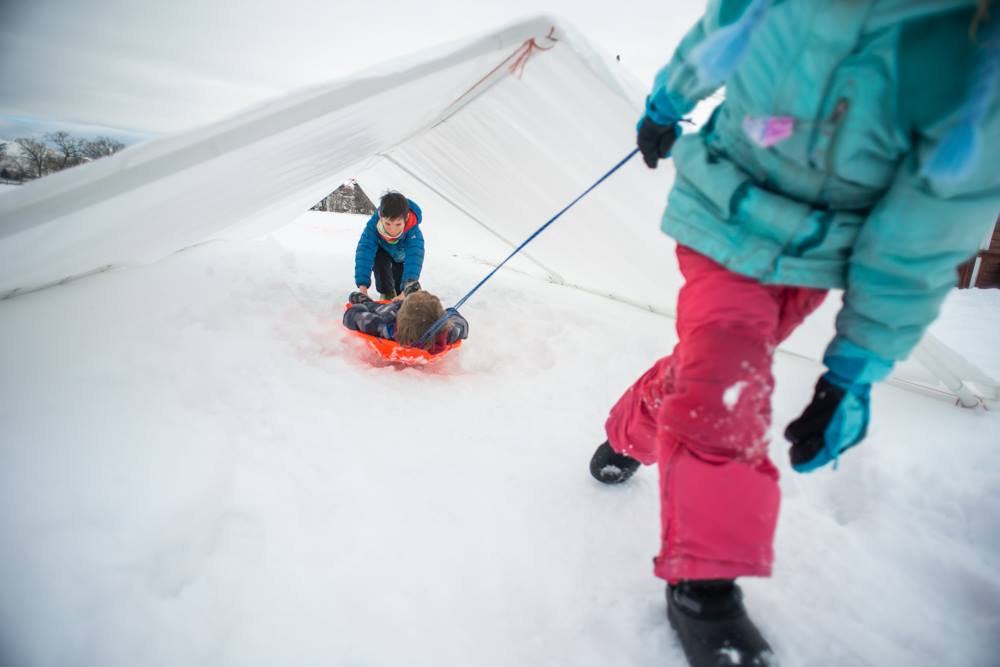 A girl in pink snow pants pulls a boy on an orange sled as they play outside in winter.
