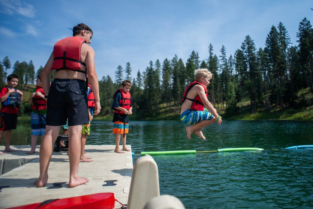 Boy Scout in lifevest cannon balls into a lake at summer camp, as other boys on dock watch.