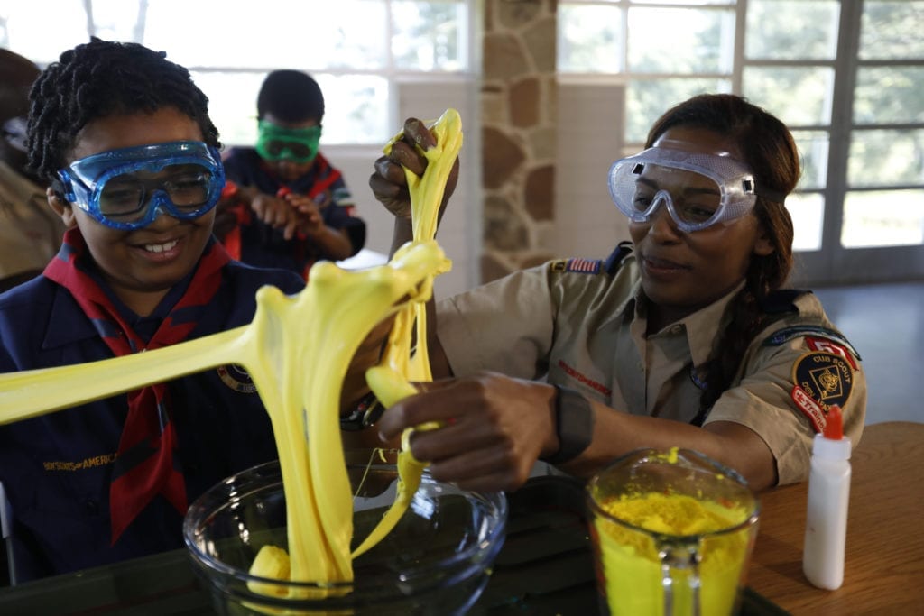 A boy and a girl wear safety goggles and stretch slime with their hands, practicing STEM skills at summer camp.