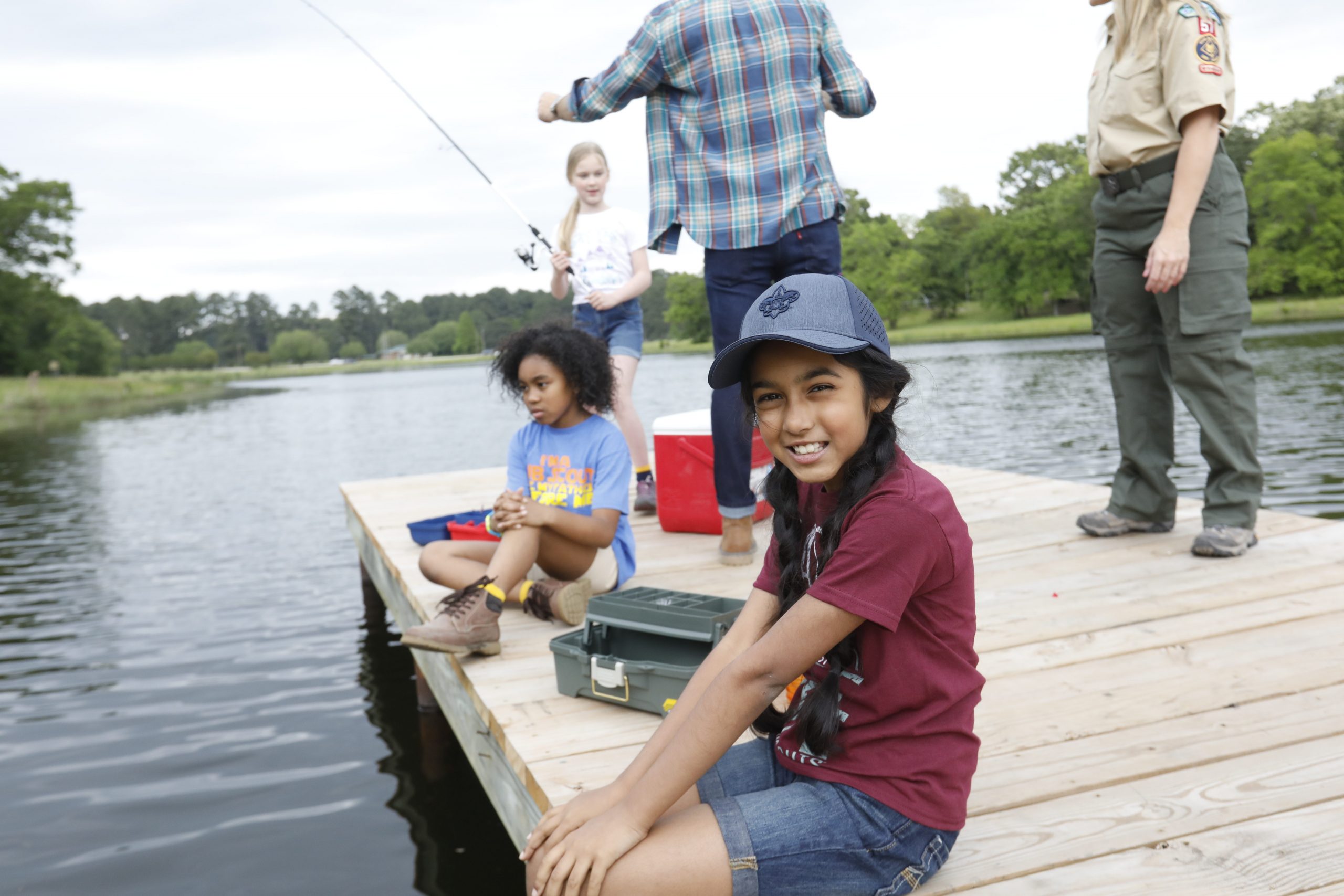 Girl wearing hat smiles as she sits on dock, fishing in lake at summer camp.