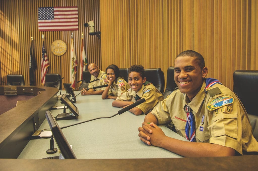 Group of older kids in Scouts BSA uniforms sit at microphones in a city council meeting.